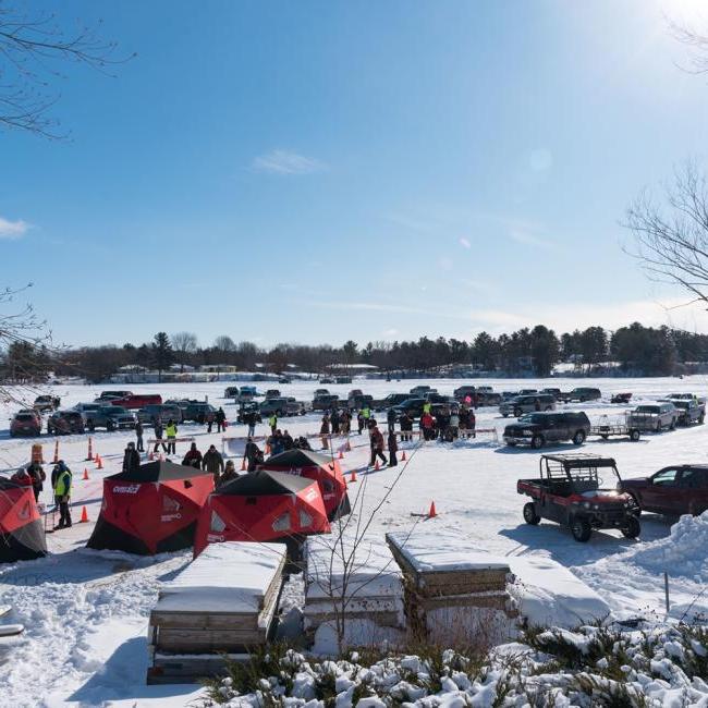 An ice fishing event on a frozen lake with participants gathered around tents and parked vehicles, enjoying winter activities in a lively, communal setting